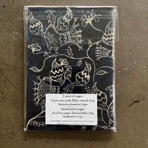 May include: A notebook with a dark blue cover featuring a white peacock and floral design. The notebook is packaged in clear plastic. The text on the label reads "Notebook 64 pages, Acid free paper, Natural white, 90g, Handbound in 7 stages."