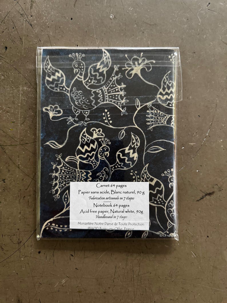 May include: A notebook with a dark blue cover featuring a white peacock and floral design. The notebook is packaged in clear plastic. The text on the label reads "Notebook 64 pages, Acid free paper, Natural white, 90g, Handbound in 7 stages."