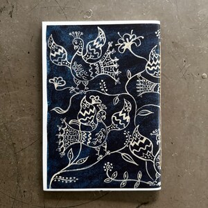 May include: A rectangular, navy blue notebook with a white border. The cover features a detailed white illustration of stylized birds, flowers, and vines. The design is intricate and decorative, creating a folk art aesthetic.