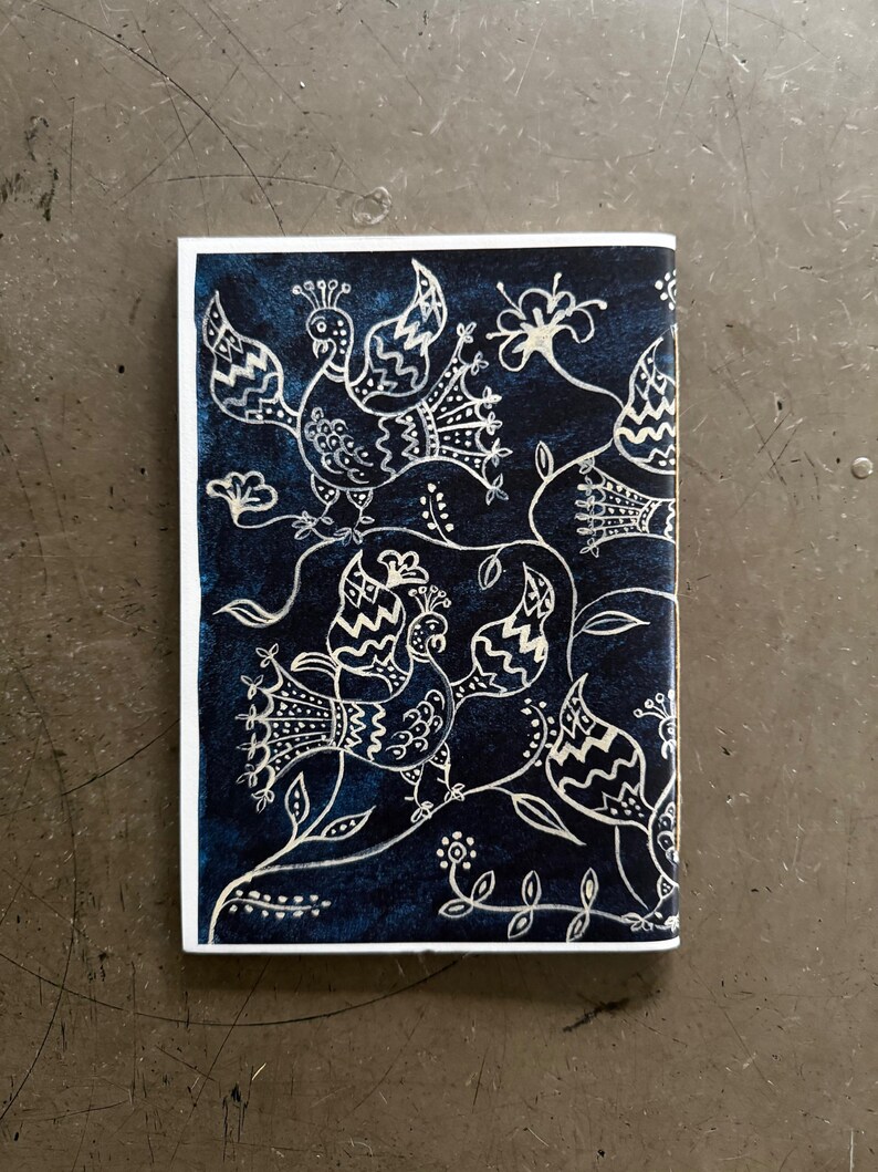 May include: A rectangular, navy blue notebook with a white border. The cover features a detailed white illustration of stylized birds, flowers, and vines. The design is intricate and decorative, creating a folk art aesthetic.