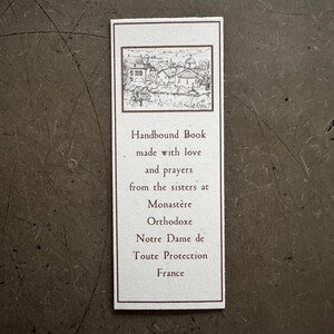 May include: A rectangular bookmark with a brown border and text. The top features a small black and white illustration of buildings. The text reads "Handbound Book made with love and prayers from the sisters at Monast&egrave;re Orthodoxe Notre Dame de Toute Protection France."