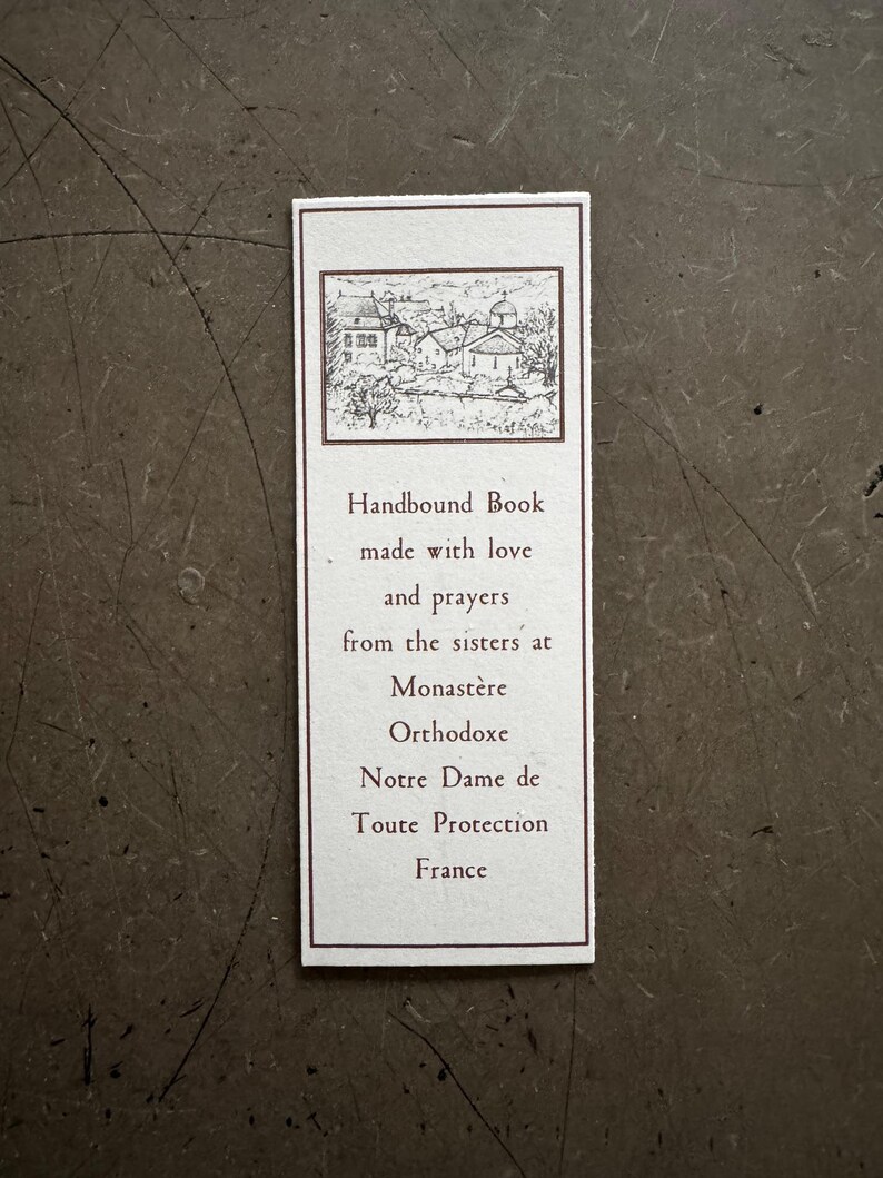 May include: A rectangular bookmark with a brown border and text. The top features a small black and white illustration of buildings. The text reads "Handbound Book made with love and prayers from the sisters at Monast&egrave;re Orthodoxe Notre Dame de Toute Protection France."
