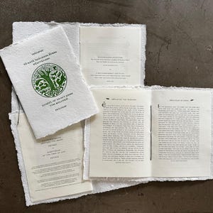 May include: A collection of handmade paper books with rough edges. The cover of the main book features green text and a circular illustration of a tree and animals. The text reads "Gospel of Saint John The Beloved Prologue".