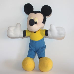 Op de afbeelding: Een Mickey Mouse knuffel met zwarte oren, een beige gezicht en een zwarte neus. Hij draagt een gele top, een blauwe broek, witte handschoenen en gele schoenen. De armen van het speelgoed zijn uitgestrekt, met duimen omhoog.