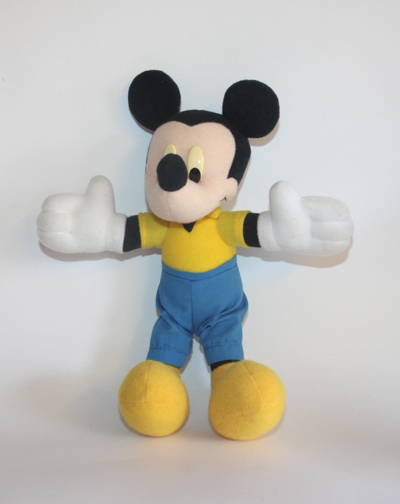 Op de afbeelding: Een Mickey Mouse knuffel met zwarte oren, een beige gezicht en een zwarte neus. Hij draagt een gele top, een blauwe broek, witte handschoenen en gele schoenen. De armen van het speelgoed zijn uitgestrekt, met duimen omhoog.