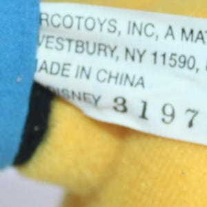 Op de afbeelding: Een close-up van een speelgoedlabel met de tekst "RCOTOYS, INC, A MATTEL CO, VESTBURY, NY 11590, USA, MADE IN CHINA, DISNEY 3197 KB". Het label is wit met zwarte tekst en bevestigd aan een geel en blauw pluche speelgoed.