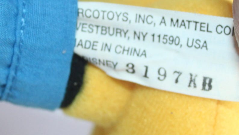 Op de afbeelding: Een close-up van een speelgoedlabel met de tekst "RCOTOYS, INC, A MATTEL CO, VESTBURY, NY 11590, USA, MADE IN CHINA, DISNEY 3197 KB". Het label is wit met zwarte tekst en bevestigd aan een geel en blauw pluche speelgoed.