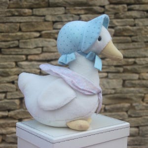 May include: A soft white goose toy, adorned with a light blue bonnet patterned with small dots and a matching bow. The goose has a yellow beak and sits on a white box, against a stone wall.
