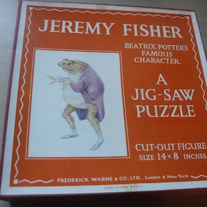 May include: A vintage jigsaw puzzle box featuring "Jeremy Fisher," a character from Beatrix Potter's stories. The box is orange with white text and a picture of a frog in a purple coat. The puzzle is 14 x 8 inches.