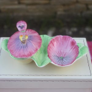 May include: A ceramic pansy-shaped dish with a lid. The pansy is painted in shades of pink and purple, with green leaves. The dish is placed on a white box.