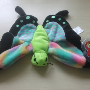 May include: A plush butterfly toy with black wings featuring green polka dots and rainbow-colored accents. The butterfly has a green body and a red heart-shaped tag.
