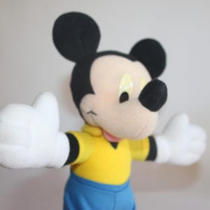 Op de afbeelding: Een Mickey Mouse knuffel met zwarte oren, een zwarte neus en witte handschoenen. Mickey draagt een geel shirt met zwarte randen en een blauwe korte broek. De armen van het speelgoed zijn uitgestrekt en het heeft een vriendelijke uitdrukking.
