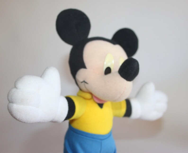 Op de afbeelding: Een Mickey Mouse knuffel met zwarte oren, een zwarte neus en witte handschoenen. Mickey draagt een geel shirt met zwarte randen en een blauwe korte broek. De armen van het speelgoed zijn uitgestrekt en het heeft een vriendelijke uitdrukking.