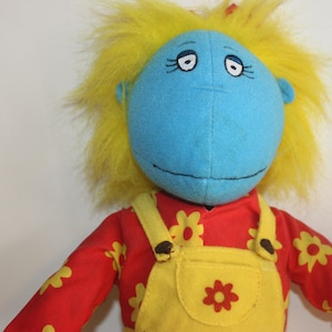 May include: A soft toy doll with a blue face, yellow hair, and a red shirt with yellow flower patterns. The doll is wearing yellow dungarees with a flower design. The doll has a smiling expression.