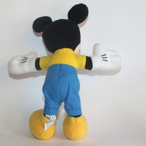 Op de afbeelding: Een pluche Mickey Mouse-speelgoed, gezien van achteren, met zwarte oren en hoofd, een geel shirt, blauwe broek, witte handschoenen en gele schoenen. De armen van het speelgoed zijn uitgestrekt, tegen een witte achtergrond.