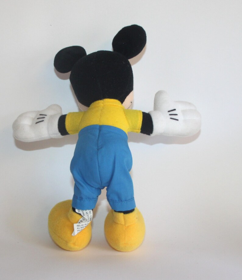 Op de afbeelding: Een pluche Mickey Mouse-speelgoed, gezien van achteren, met zwarte oren en hoofd, een geel shirt, blauwe broek, witte handschoenen en gele schoenen. De armen van het speelgoed zijn uitgestrekt, tegen een witte achtergrond.
