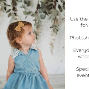 May include: A little girl wearing a light blue denim dress with a white lace collar and a gold bow in her hair. The girl is looking to the right. The background is a white wall with greenery hanging from the top.