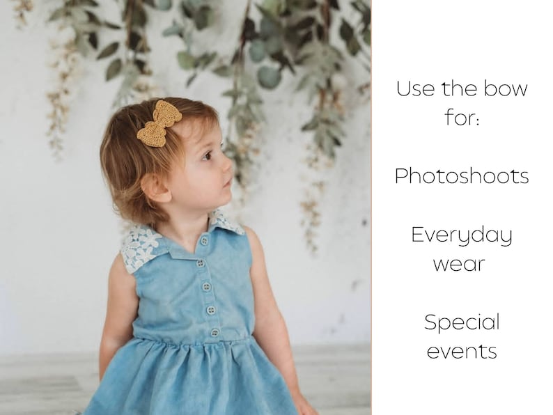 May include: A little girl wearing a light blue denim dress with a white lace collar and a gold bow in her hair. The girl is looking to the right. The background is a white wall with greenery hanging from the top.