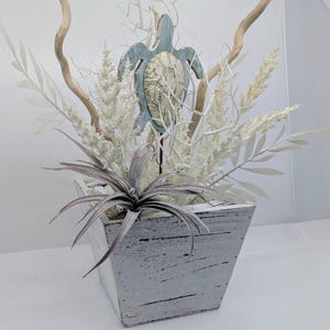 May include: A decorative arrangement in a white distressed square wooden planter featuring a central sea turtle cutout with a textured shell, surrounded by white faux floral sprigs, thin white branches, and a spiky grey-toned air plant.