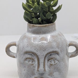 May include: A ceramic face planter with a distressed white finish and two small handles. The planter features a sculpted face with closed eyes and a smiling mouth. It is filled with a green succulent plant.