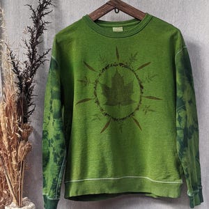 Pode incluir: Camisola verde com gola redonda e mangas compridas. A frente apresenta um design circular com uma folha no centro e detalhes de folhas e caules. As mangas têm um efeito tie-dye em tons de verde.