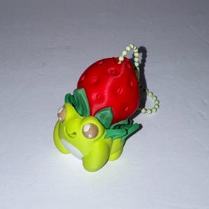 May include: A novelty keychain showing a green frog with a red strawberry on its back. The frog has large eyes and green leaves. The keychain has a beaded chain attached.