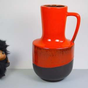 Könnte beinhalten: Eine große, orangefarbene und schwarze Keramikvase mit Henkel. Die Vase hat eine glänzende Oberfläche und eine abgerundete Form.