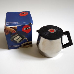 May include: A silver creamer with a black lid and handle. The creamer has a circular design on the lid. The creamer is in a box with the word "QUIST" printed on it. The box is blue and white with German text.