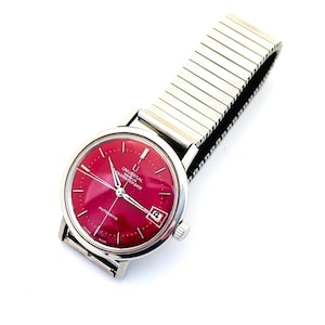 Può includere: Un orologio da polso vintage con quadrante rosso acceso e cassa e cinturino color argento. Il quadrante presenta la scritta "UNIVERSAL GENEVE" e "AUTOMATIC". La data è visualizzata alle ore 3. Il cinturino è di tipo espandibile, color argento.