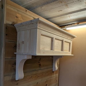 May include: A wooden shelf unit with a decorative top and three recessed panels. The shelf is supported by two curved brackets. The wood has a natural, unfinished appearance, and is mounted on a wooden wall.