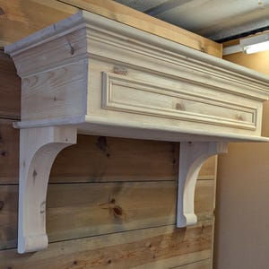 May include: An unfinished wooden mantelpiece with decorative moulding and two curved support brackets. The mantelpiece is mounted on a wooden wall, showcasing a rustic design.