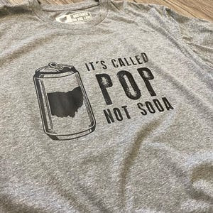 May include: Heather gray t-shirt with a graphic of a soda can featuring the Ohio state outline. The text reads "IT'S CALLED POP NOT SODA". The shirt also has the text "MISTAKES ON THE LAKE" on the tag.
