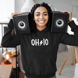 Könnte beinhalten: Ein schwarzer Hoodie mit einem weißen Aufdruck, der "OHIO" mit einem Blitz zeigt. Das Model trägt den Hoodie und hält zwei schwarze Lautsprecher.