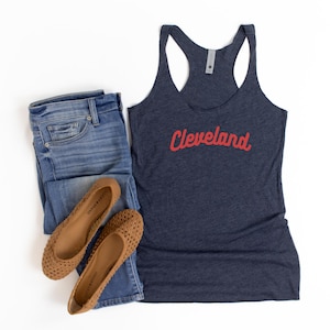 Puede incluir: Una camiseta sin mangas azul marino con la palabra "Cleveland" en escritura roja, combinada con vaqueros azules y bailarinas marrones. El atuendo informal está dispuesto sobre un fondo blanco.