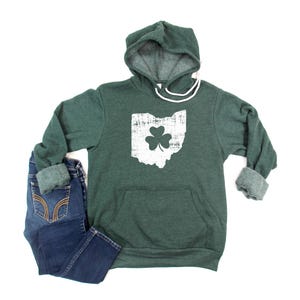 Puede incluir: Una sudadera con capucha verde con un contorno blanco del estado de Ohio y un trébol en su interior. La sudadera tiene un bolsillo canguro.