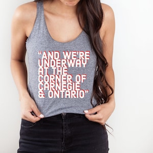 May include: Gray tank top with the text "AND WE'RE UNDERWAY AT THE CORNER OF CARNEGIE & ONTARIO" in red and white. The top is a scoop neck style and is made of a soft material. The text is centered on the front of the tank top.