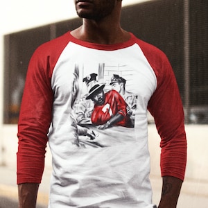 May include: A white and red baseball t-shirt with a black and white illustration of a man in a red shirt being arrested. The illustration is on the front of the shirt.