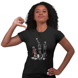 May include: Black t-shirt with a graphic of five figures, including a football player kneeling, and two figures with raised fists. The figures are in black and white, except for the football player who is wearing a red and white jersey.