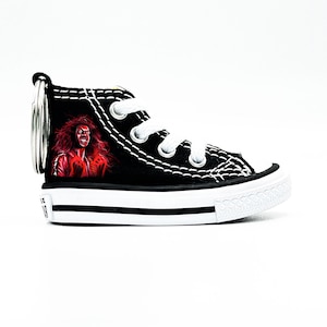 May include: Black canvas high-top sneaker keychain with a red and black illustration of a person with long hair. The keychain has a silver metal ring.