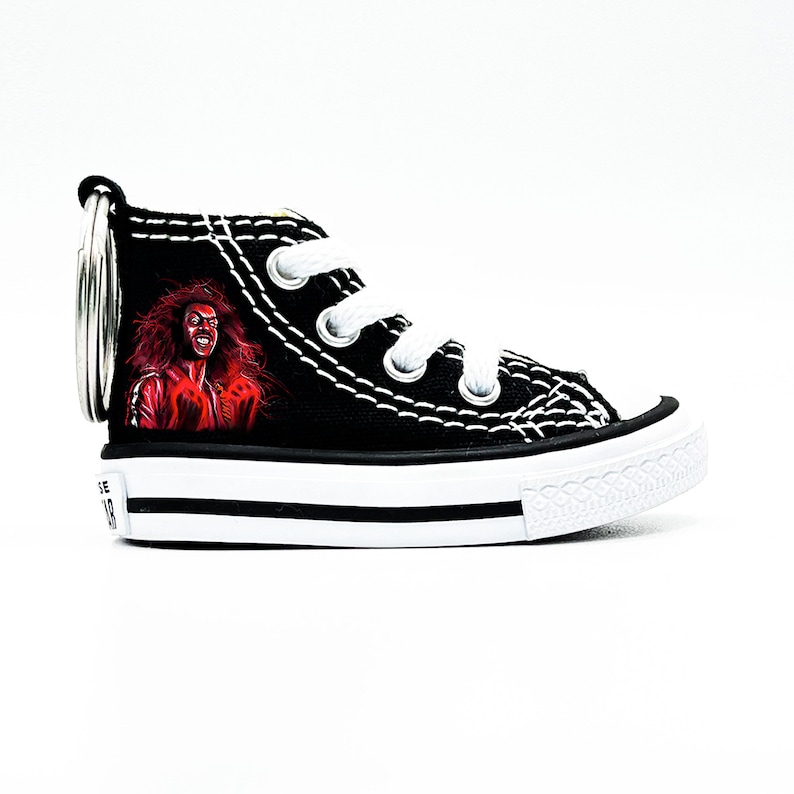 May include: Black canvas high-top sneaker keychain with a red and black illustration of a person with long hair. The keychain has a silver metal ring.