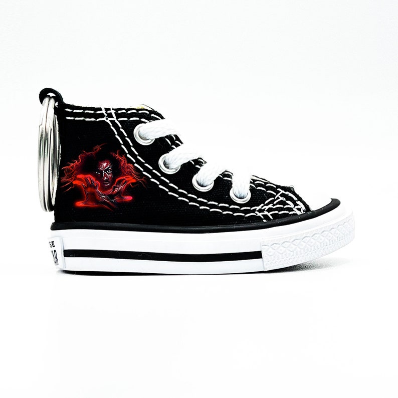 May include: Black canvas high-top sneaker keychain with a red and black graphic of a person's face. The keychain has a silver metal ring.
