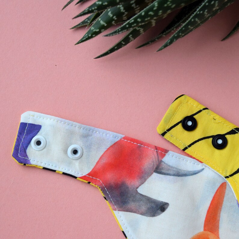 May include: A fabric bib featuring a colourful graphic print of animals on a white background. The bib has two white plastic snap fasteners on one side and two black plastic snap fasteners on the other side, with yellow fabric trim.