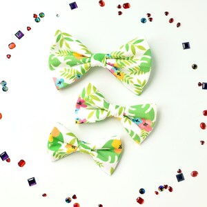 May include: Three bow ties of varying sizes featuring a white background with a vibrant tropical pattern of green palm leaves and small multi-coloured flowers in shades of pink, yellow, and blue, surrounded by scattered loose colourful rhinestones.