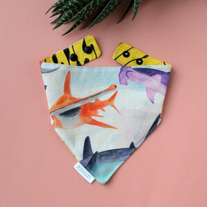 May include: A triangular bandana featuring a watercolour print of orange and purple hammerhead sharks on a light background. The reverse side is bright yellow with a black abstract pattern. It includes black snap buttons and a white tag reading CRAZYCOOLCAT.