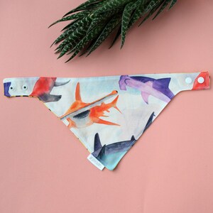 May include: A triangular pet bandana featuring a watercolour print of various sharks in shades of orange, purple, and grey on a light background. The accessory includes two white plastic snap buttons on each end and a small white fabric tag.