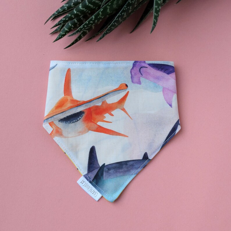 May include: A triangular pet bandana featuring a watercolour print of various sharks, including orange and purple hammerhead sharks, on a light blue background. A small white fabric tag with the text CRAZYCOOLCAT is sewn onto the bottom edge.