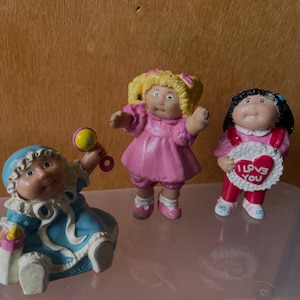 May include: Three small Cabbage Patch Kids figurines. One is a blue-clad baby holding a yellow rattle. The second is a blonde child in a pink dress. The third is a dark-haired child holding a white heart-shaped sign with the text I LOVE YOU.