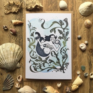Puede incluir: Una tarjeta de felicitaci&oacute;n con una ilustraci&oacute;n de lin&oacute;leo de una sirena con un sujetador de concha y un espejo, rodeada de algas marinas y un fondo azul acuarela.