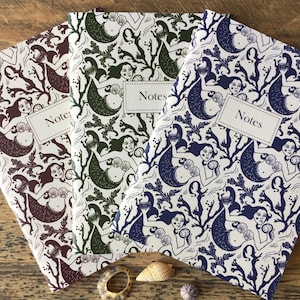 May include: Three notebooks featuring a mermaid design. The notebooks are white with a green, brown, and blue mermaid pattern. The notebooks have the word "Notes" printed on the front.