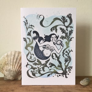 Puede incluir: Una ilustraci&oacute;n en blanco y negro de una sirena con el pelo largo, sosteniendo un espejo, rodeada de algas y burbujas. El fondo es una acuarela de color azul claro.